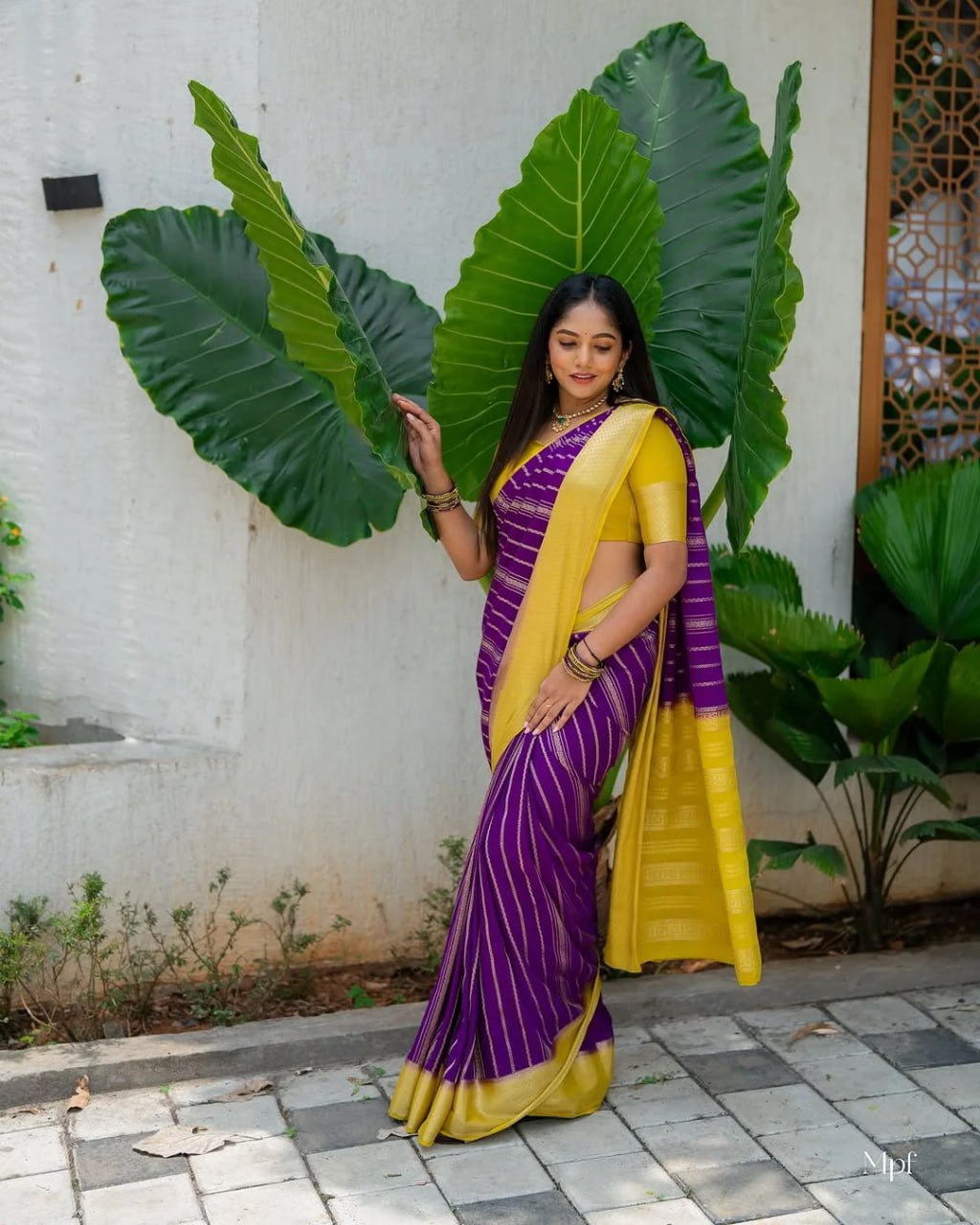 Woman in a purple and yellow saree standing outdoors with large green leaves.